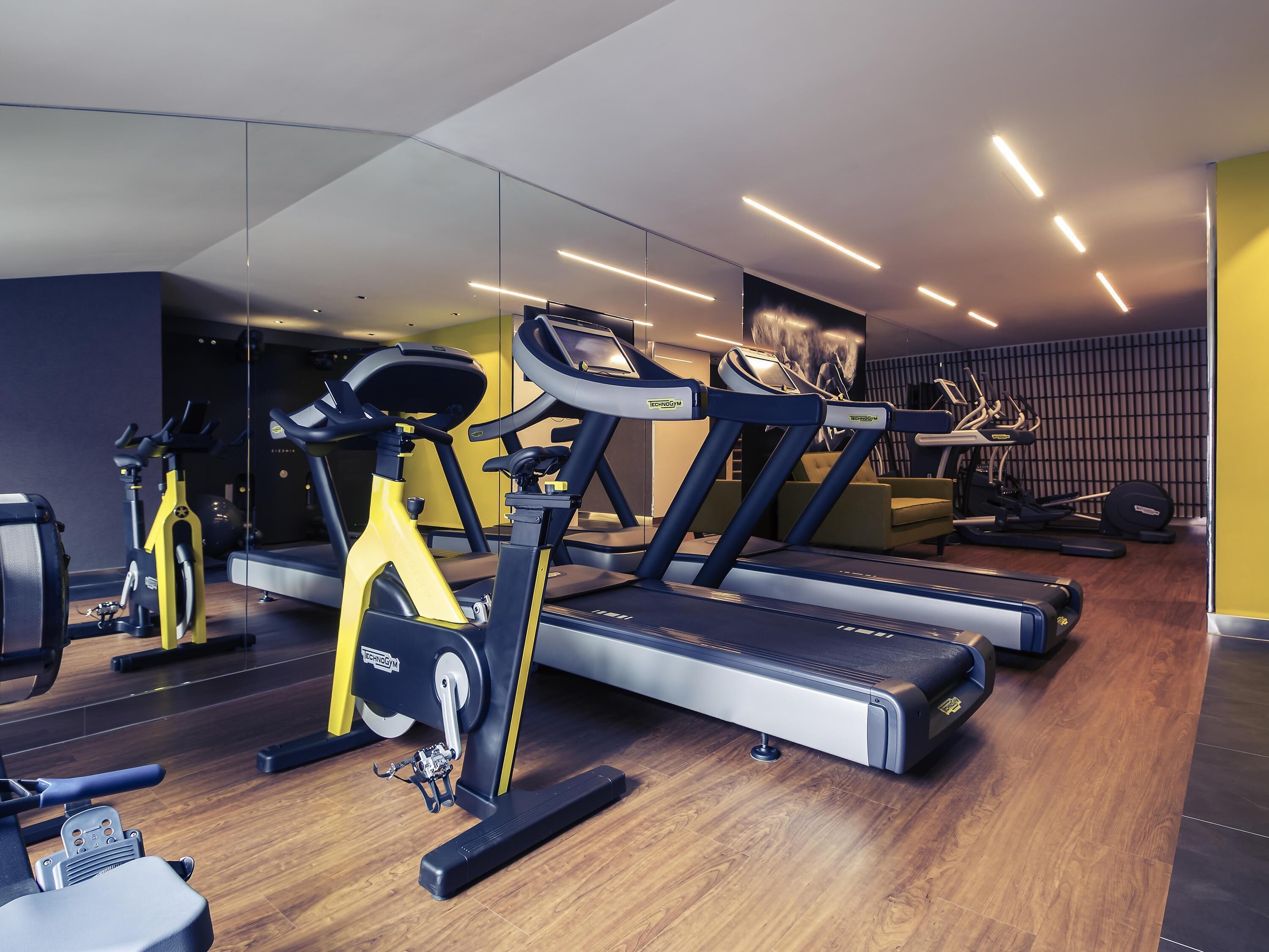 Un gimnasio con equipamiento de ejercicio, incluyendo cintas de correr y una elíptica, con suelo de madera y un espejo grande.