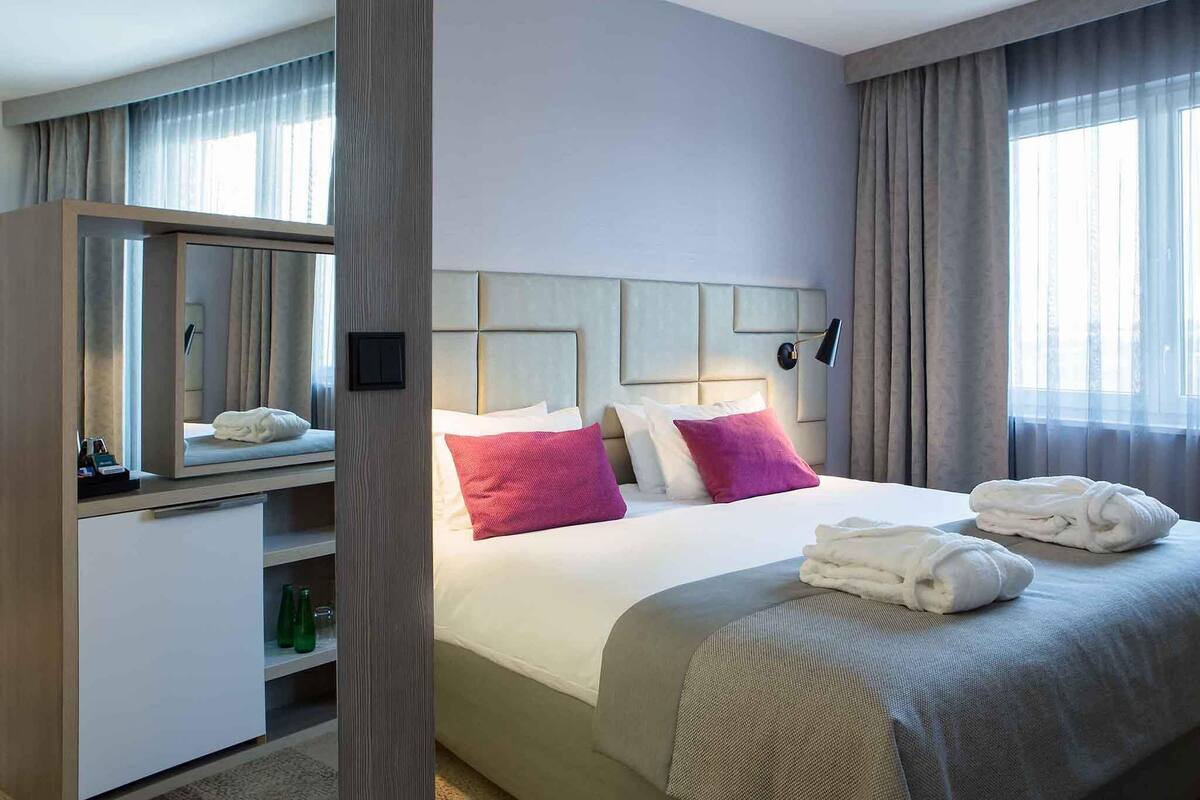Une chambre d’hôtel avec un lit, une table de chevet, un miroir et une fenêtre avec des rideaux.