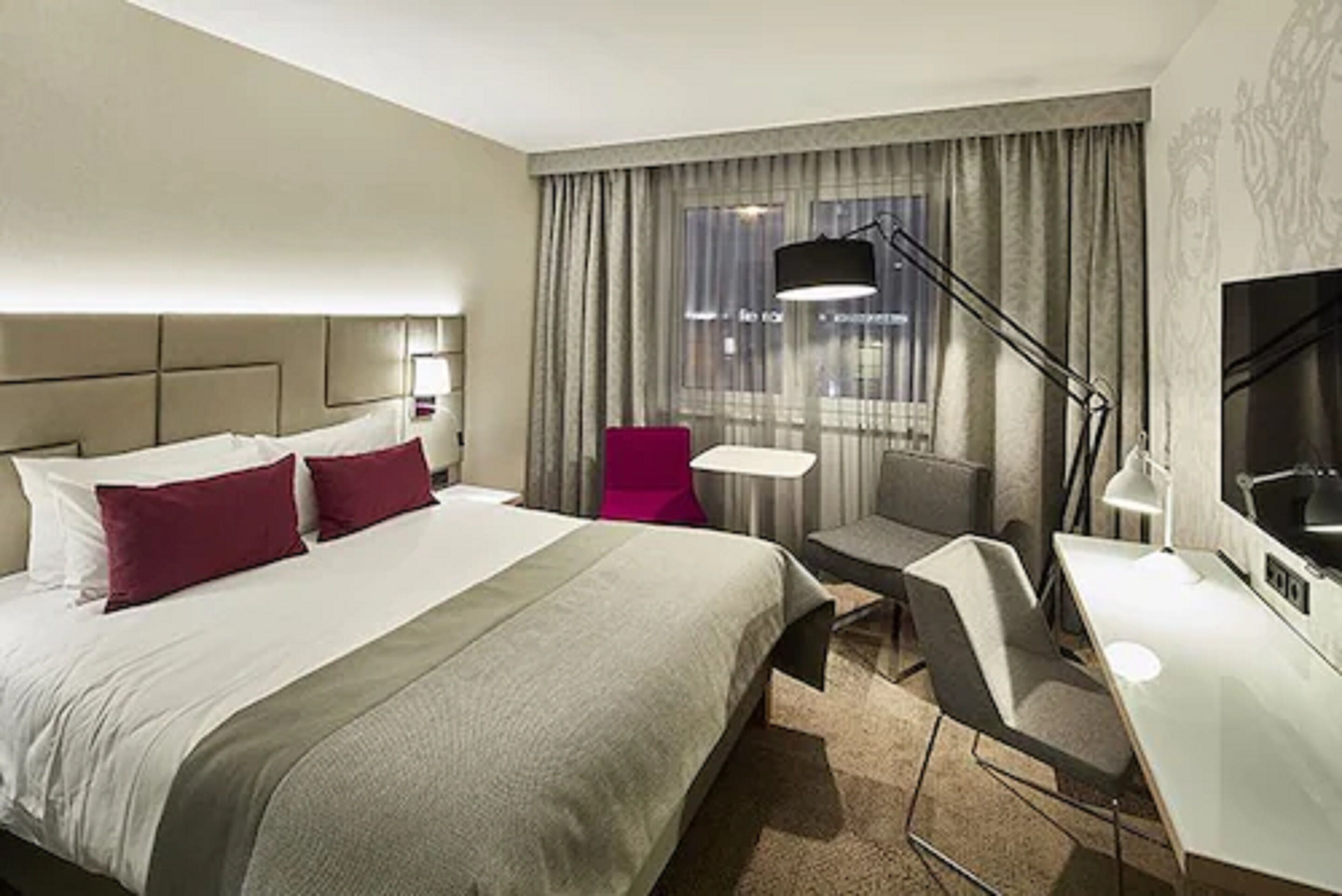 Ett hotellrum med en stor säng, ett skrivbord, en stol, en tv och ett fönster med gardiner.