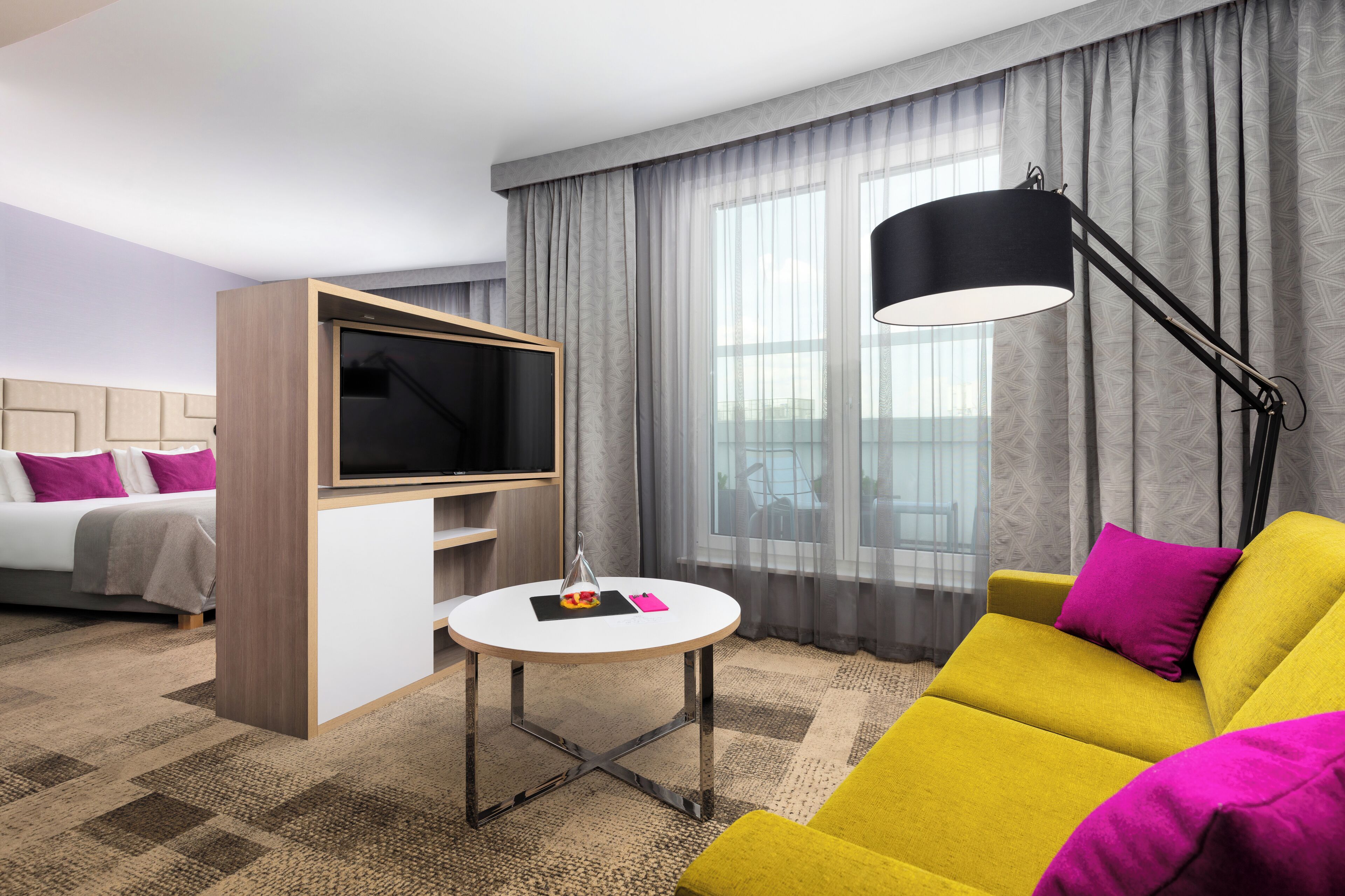 A modern hotel room with a sofa, a TV, a lamp, and a view of the city through large windows.