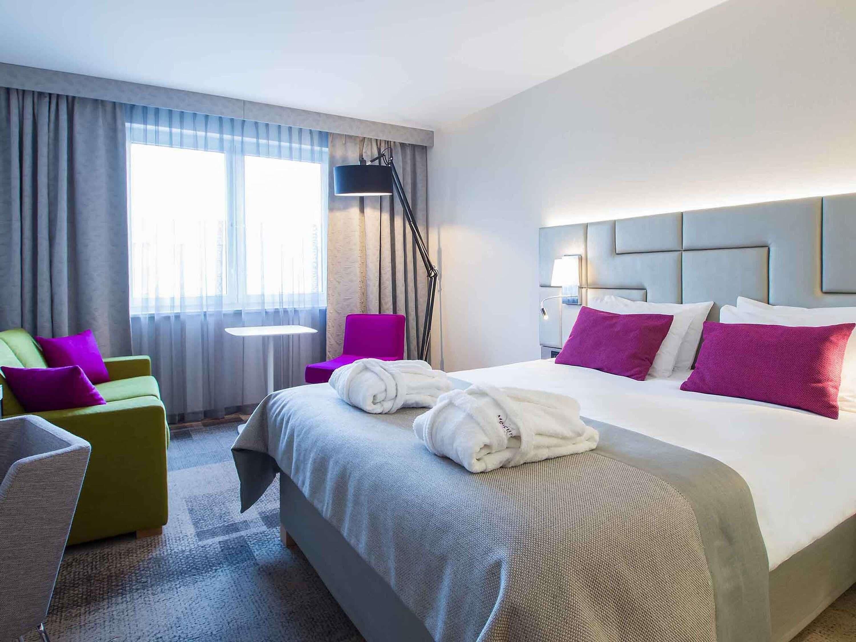A hotel room with a bed, a desk, a chair, a lamp, and a window.