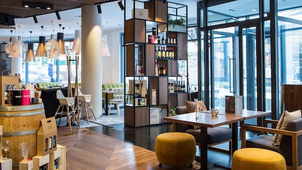 A modern interior with wooden flooring, a bar area with wine bottles, and a seating area with yellow ottomans. - Mercure Krakow Stare Miasto (Old Town) (Kraków)