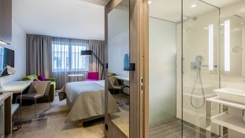 A hotel room with a bed, desk, chair, and a glass-enclosed shower area.