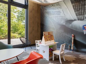 A room with a large window, a white table, orange and white chairs, a wooden desk with a pegboard, and a basketball hoop sticker on the wall. - ibis Saint Quentin En Yvelines Velodrome (Montigny-le-Bretonneux)