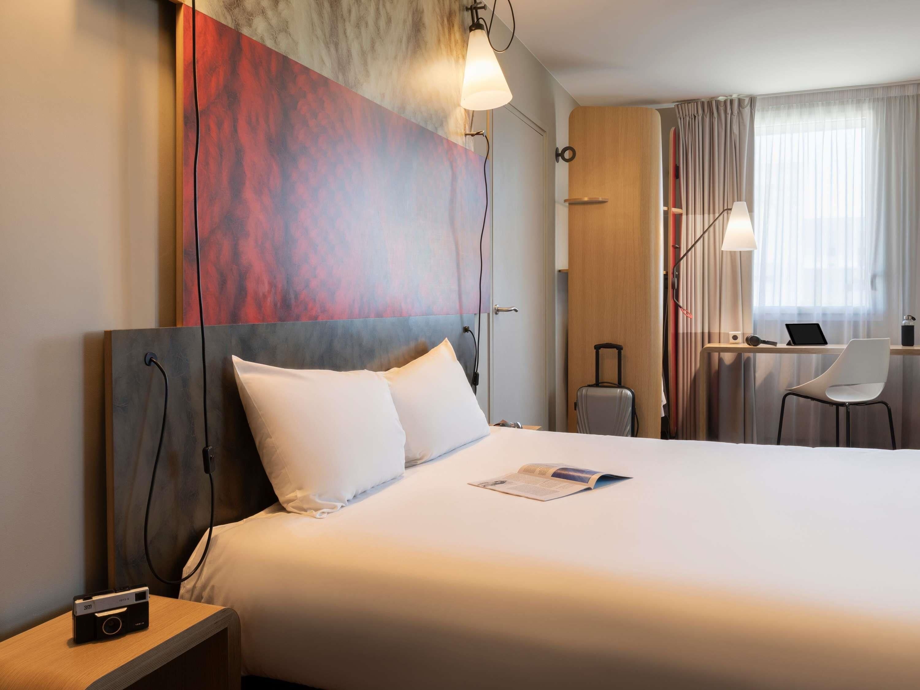 A hotel room with a bed, a desk with a laptop, a chair, a suitcase, and a wall art piece.