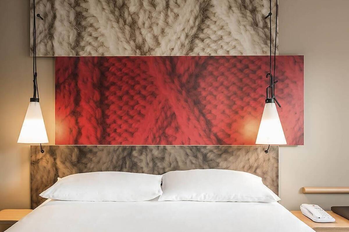 Une chambre d’hôtel avec un lit, deux lampes suspendues, un tableau abstrait rouge au-dessus du lit et un mur à la texture évoquant le marbre.