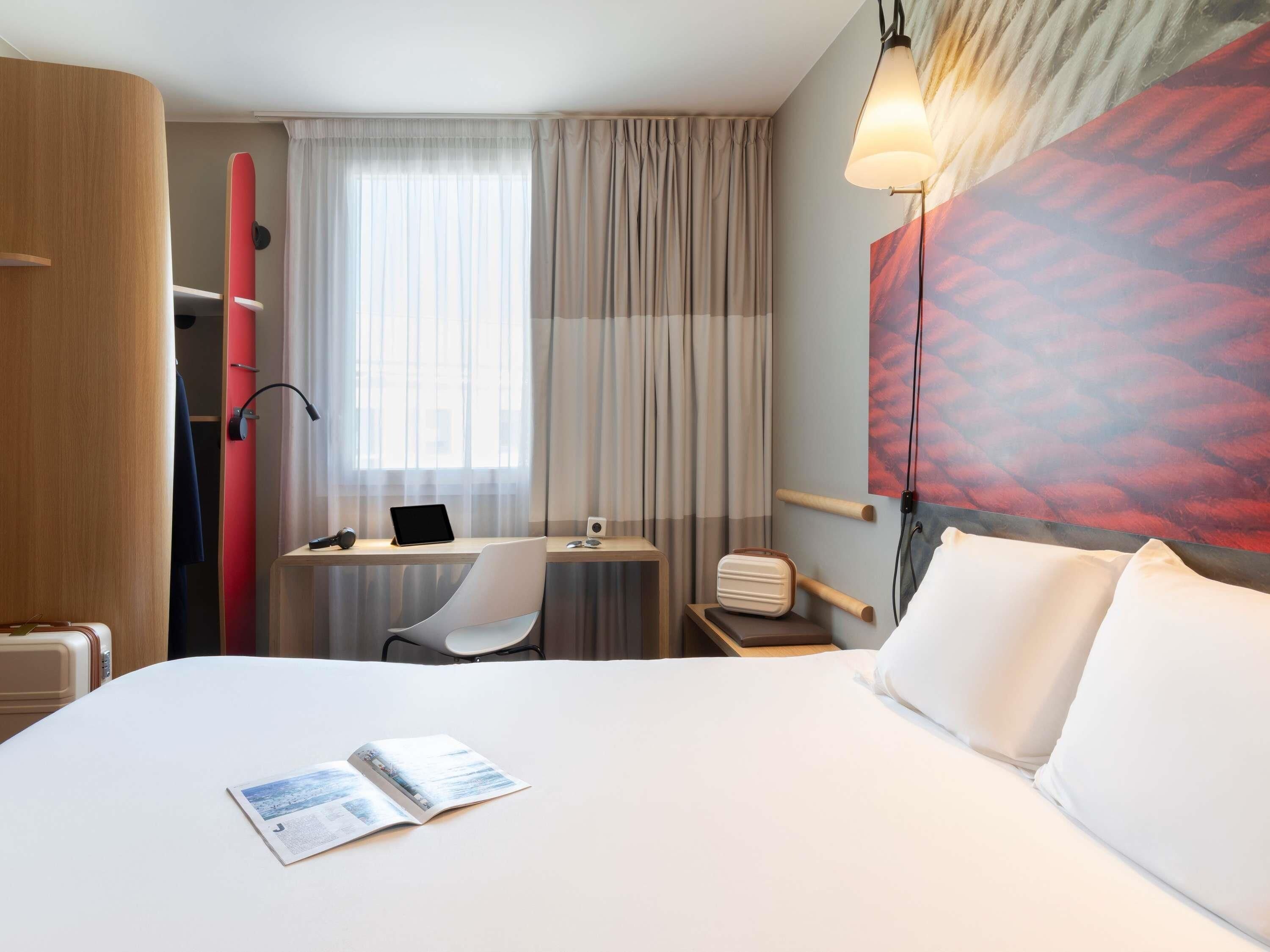 A hotel room with a bed, a desk with a laptop, a chair, a small table with a speaker, and a large window with curtains.