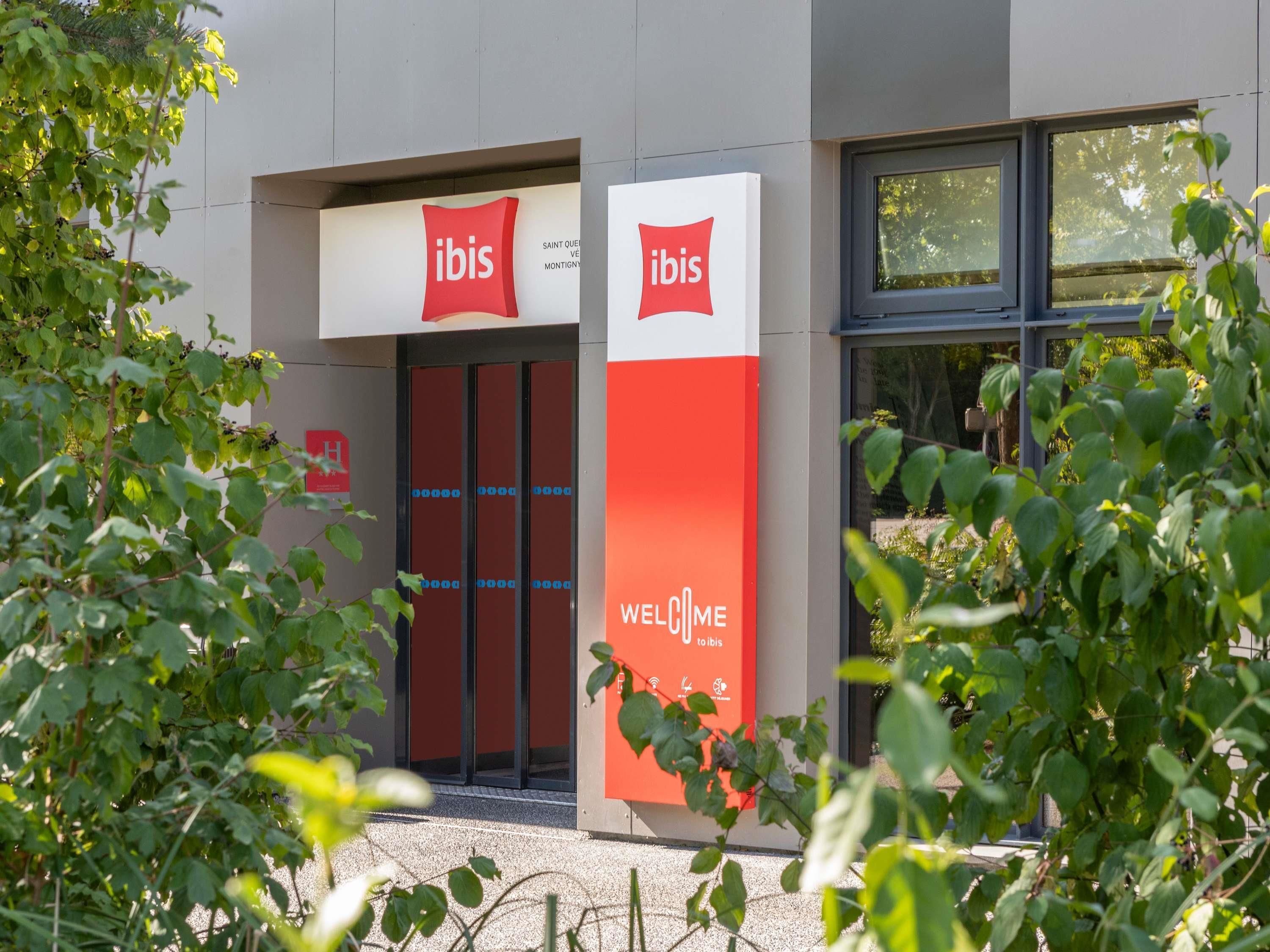 L’entrée d’un hôtel Ibis, avec une façade aux couleurs rouge et blanche, des portes en verre et une enseigne accueillant les clients.