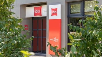 L’entrée d’un hôtel Ibis, avec une façade aux couleurs rouge et blanche, des portes en verre et une enseigne accueillant les clients.