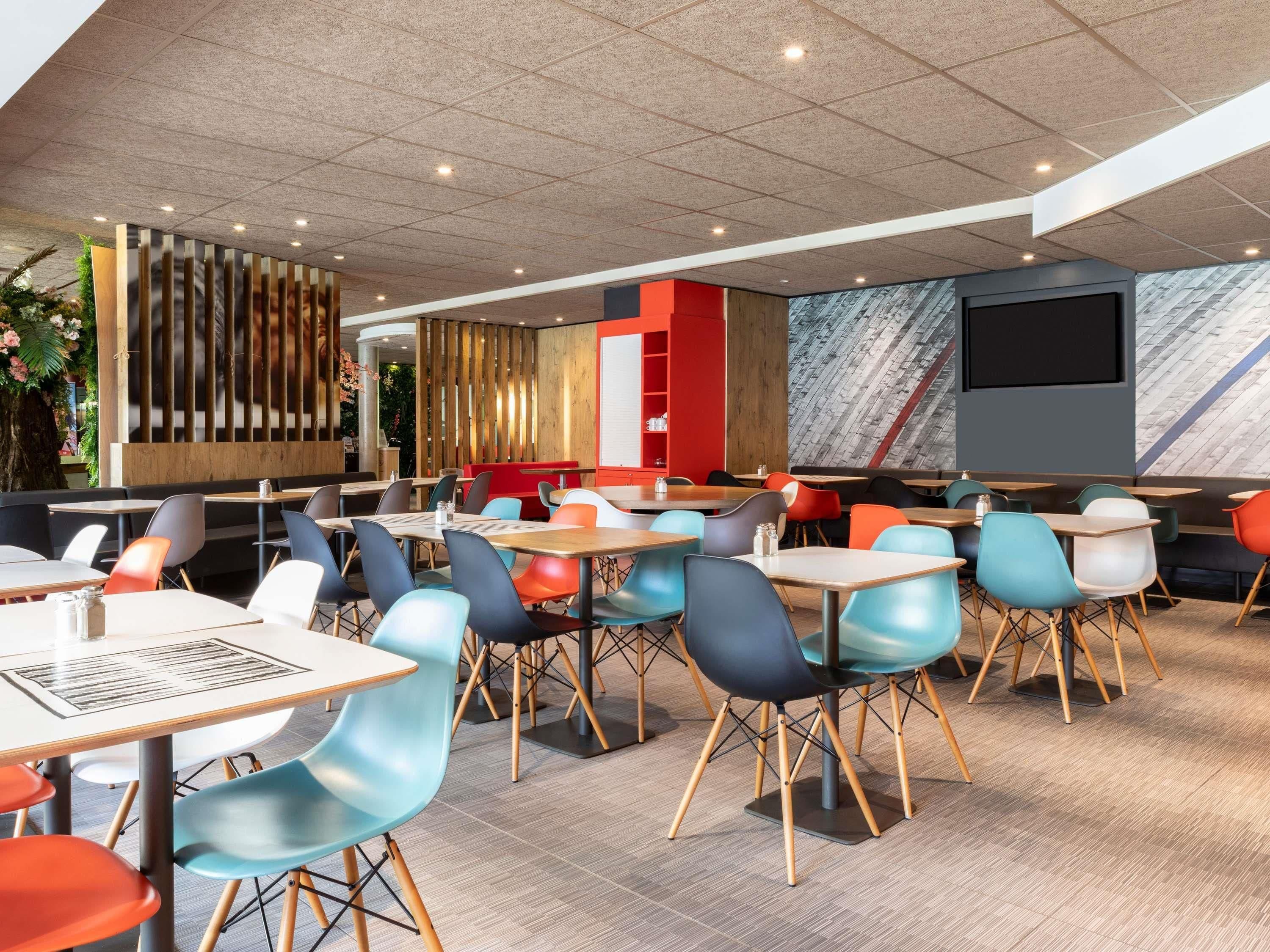A modern restaurant interior with multiple tables and chairs, a wooden accent wall, and a large screen.
