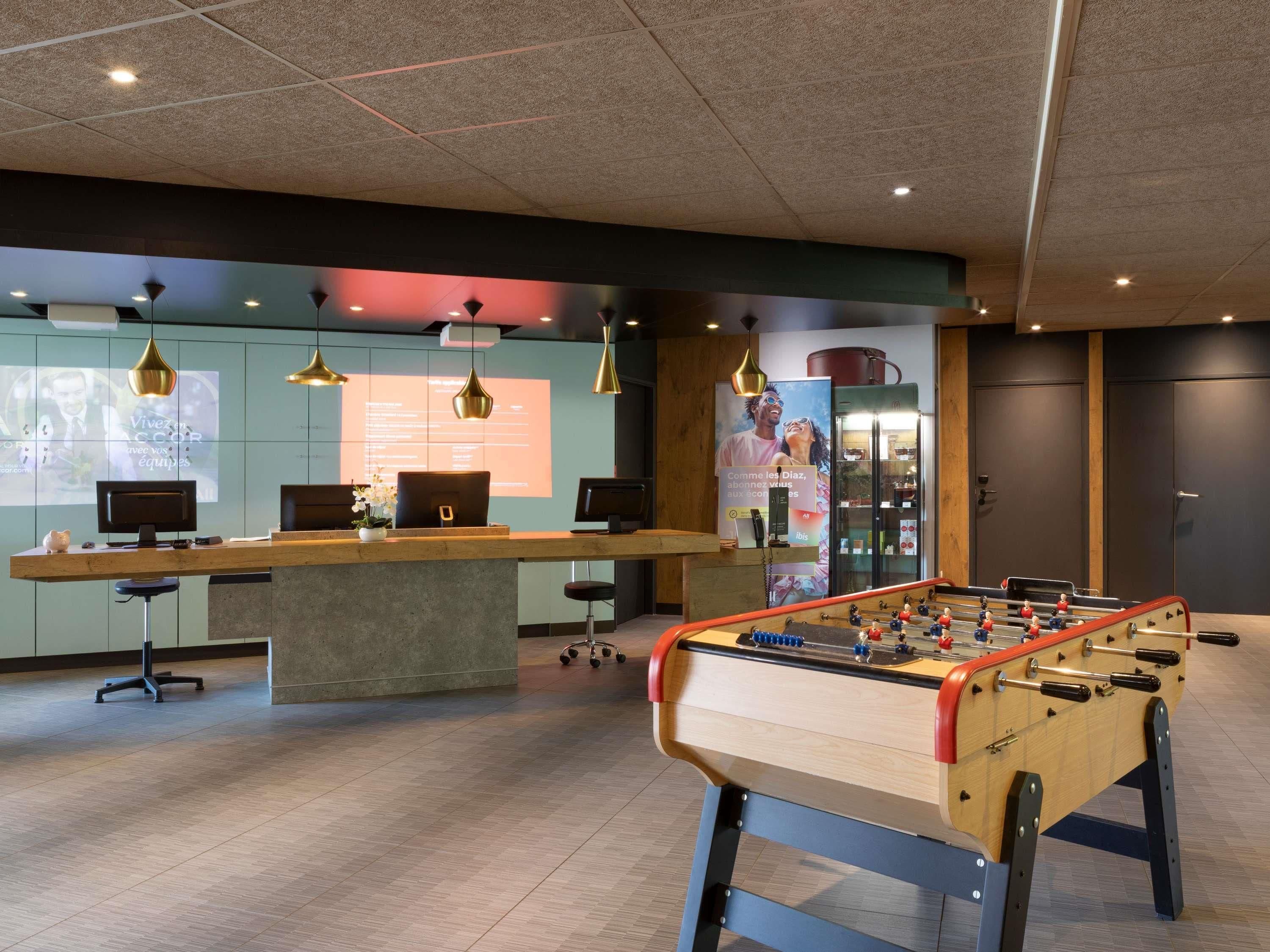 Espai interior modern amb taula de futbolí, taulell de recepció i una pantalla de projecció que mostra text.