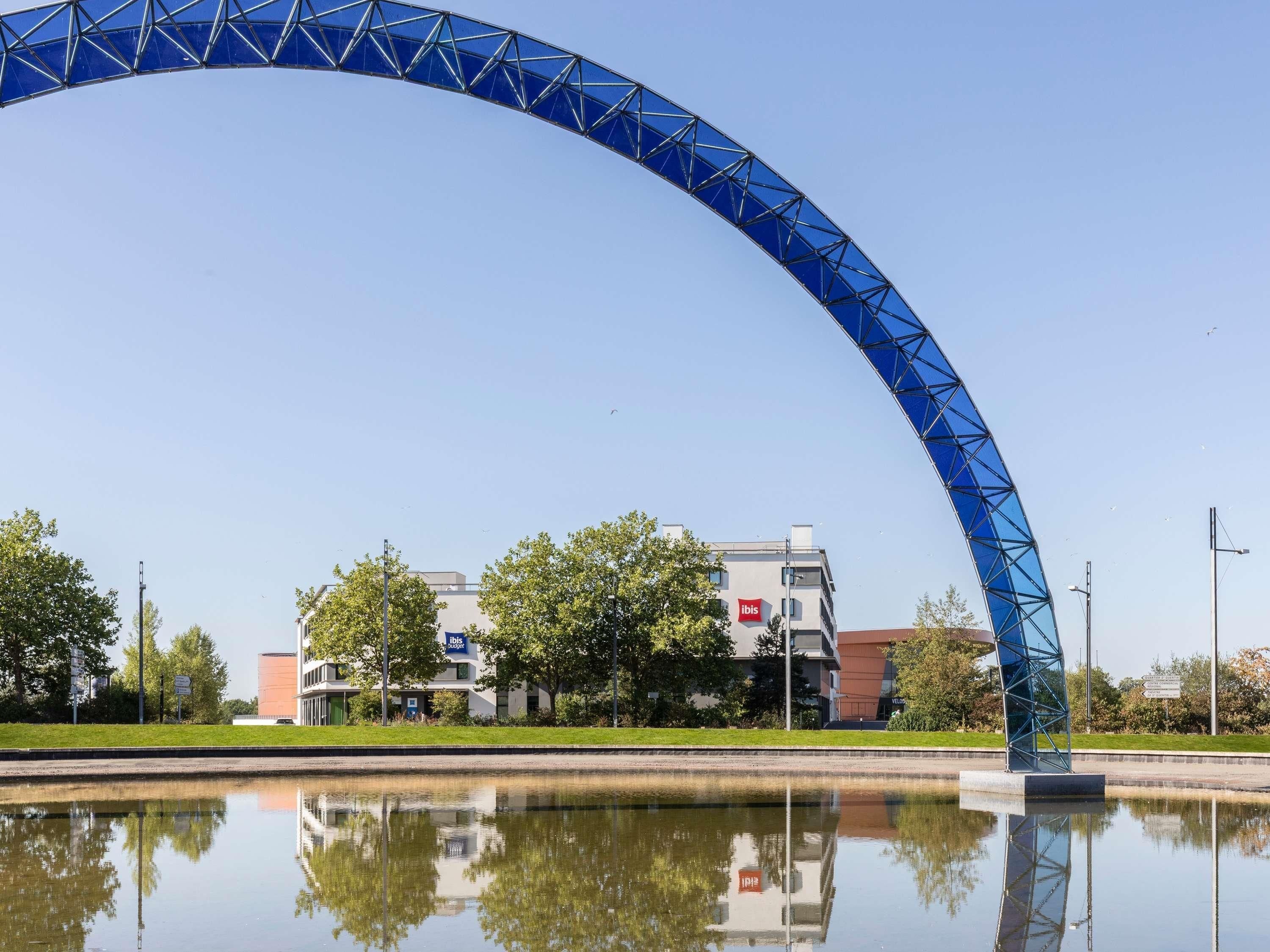 A blue arched structure over a body of water with a reflection, a building with an 'ibis' sign, and trees in the background.