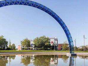 Une structure bleue en forme d’arche surplombe une étendue d’eau dont le reflet s’y projette, un bâtiment portant une enseigne avec le mot « ibis », et des arbres en arrière-plan.