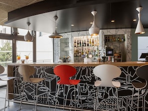 A bar area with a black and white mural, a variety of bottles, and modern pendant lighting. - ibis Saint Quentin En Yvelines Velodrome (Montigny-le-Bretonneux)