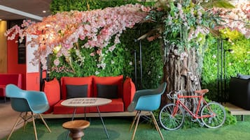 Ein Sitzbereich mit einem roten Sofa, blauen Stühlen und einem Holztisch. Ein rotes Fahrrad steht neben einem Baum.