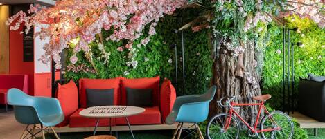 A seating area with a red sofa, blue chairs, and a wooden table. A red bicycle is parked next to a tree.