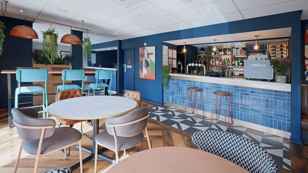 A modern bar area with a blue tiled backsplash, a bar counter, and a variety of seating options including round tables and bar stools. - Travelodge Weston-super-mare (West Malling)