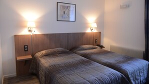 A hotel room with two beds, a wooden headboard, bedside lamps, a framed picture on the wall, and a radiator. - Hotel Du Lion (Vesoul)
