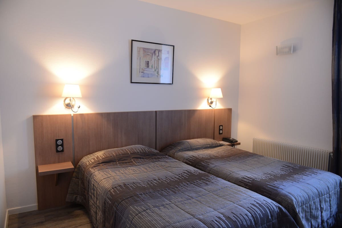 Hotellihuone, jossa on kaksi sänkyä, puinen pääty, yöpöytävalaisimet, seinällä oleva kehystetty kuva ja patteri.
