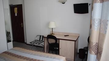 Una habitación pequeña y sencilla con cama, escritorio, silla, lámpara, televisor y puerta.