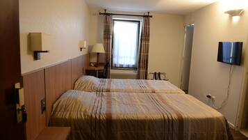 Quarto de hotel com duas camas, uma mesa de cabeceira, uma lâmpada, uma televisão e uma janela com cortinas.