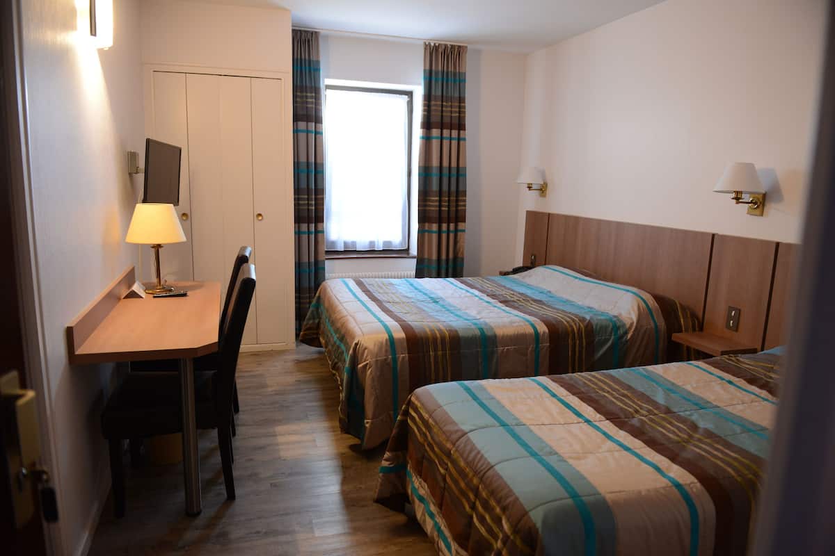 Hotellihuone, jossa on kaksi sänkyä, työpöytä lampun kanssa, tuoli ja ikkuna, jossa on verhot.