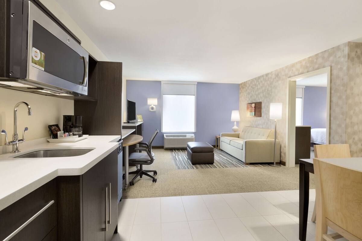 A modern kitchen with a built-in microwave, a sink, and a coffee maker. There is a seating area with a sofa and a table, and a window providing natural light.