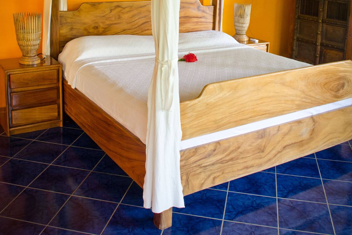 Un lit avec un cadre en bois et une literie blanche, une table de chevet avec un vase, et une porte en bois dans une pièce aux murs orange et au sol carrelé de bleu.