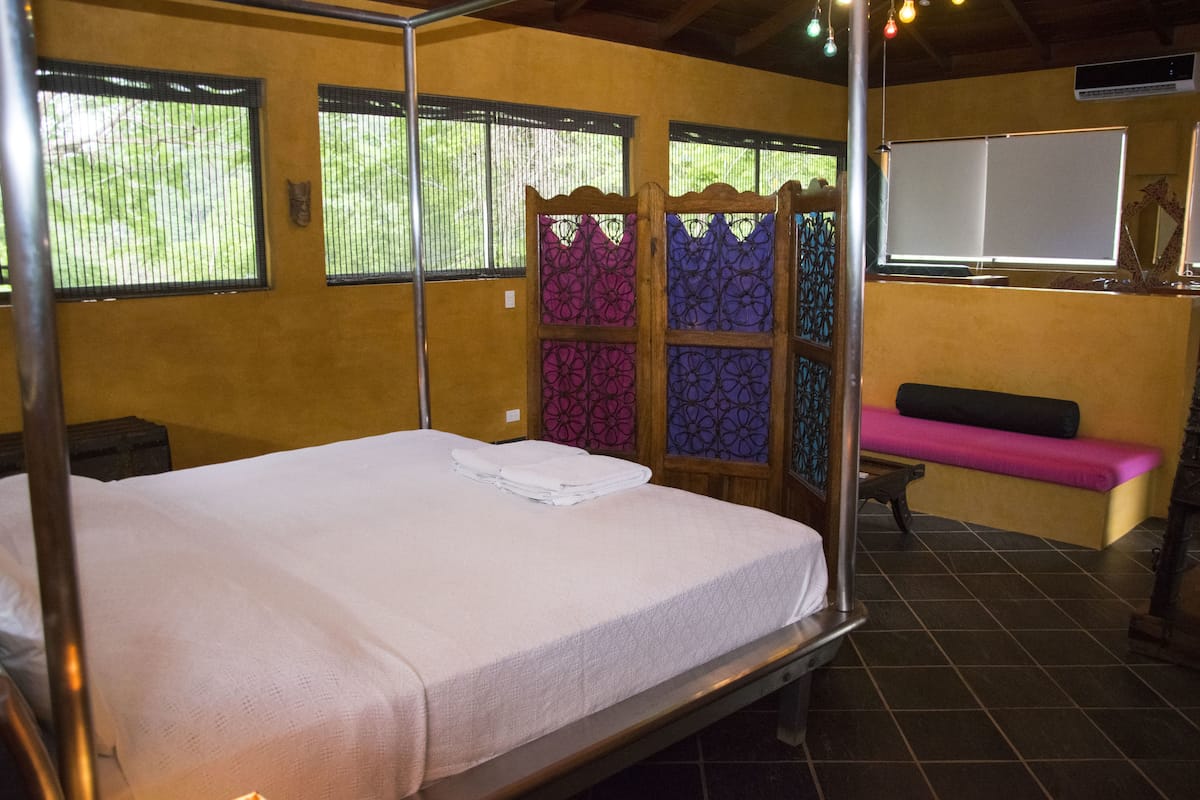 Une chambre avec un lit simple, un cadre en métal, un banc et une fenêtre donnant sur un paysage verdoyant.