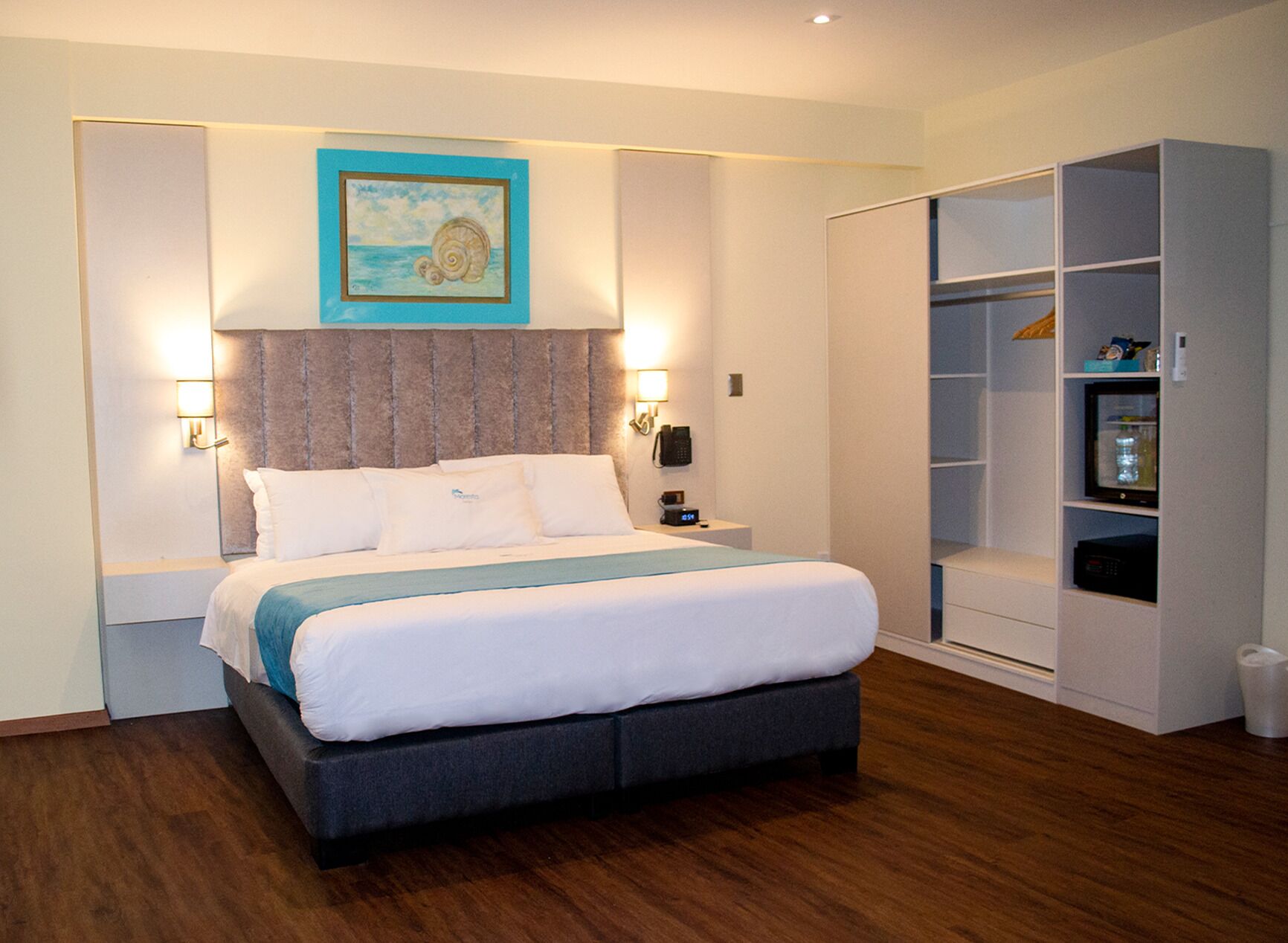 Habitación de hotel impecable con cama, lámparas de noche, un cuadro, armario y una mesita con un reloj.