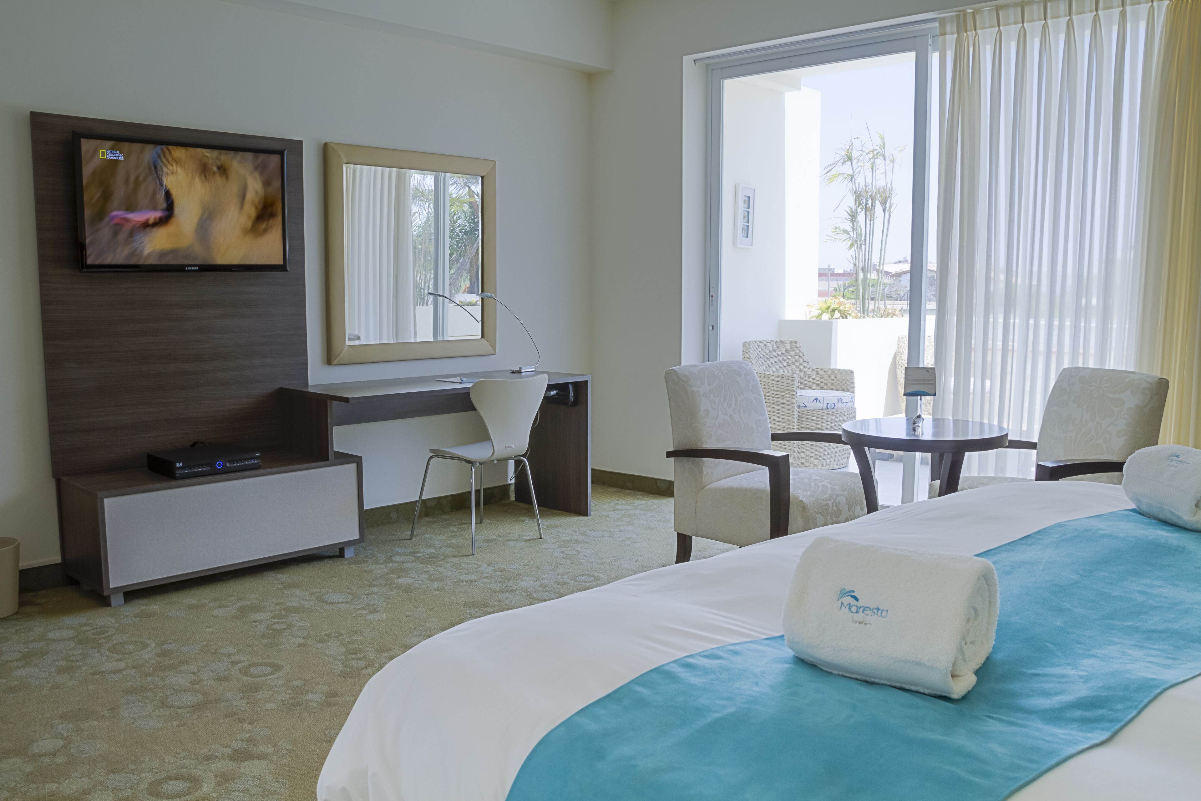 A hotel room with a large bed, a desk, a TV, and a balcony with chairs.