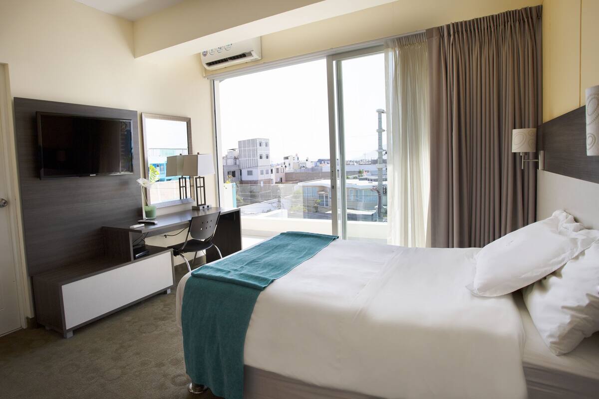 A hotel room with a bed, a desk, a TV, and a view of the city through large windows.