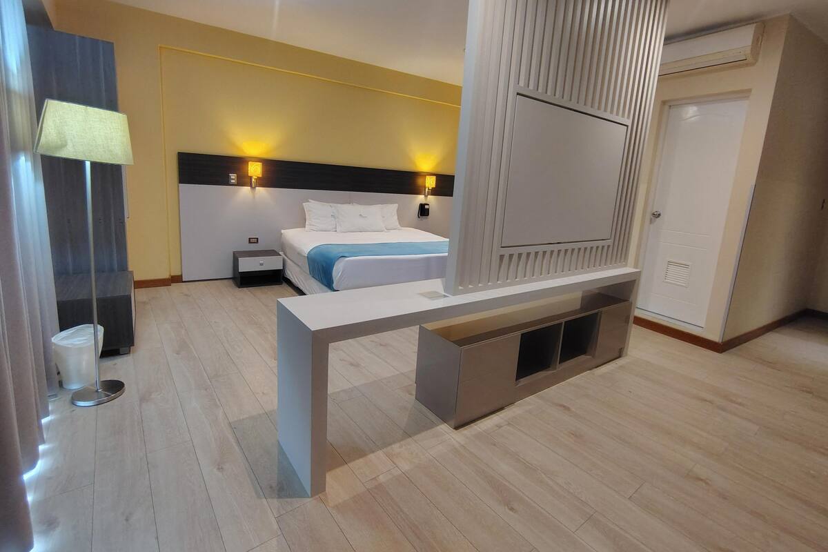 A modern hotel room with a bed, bedside tables, a TV, and a desk.