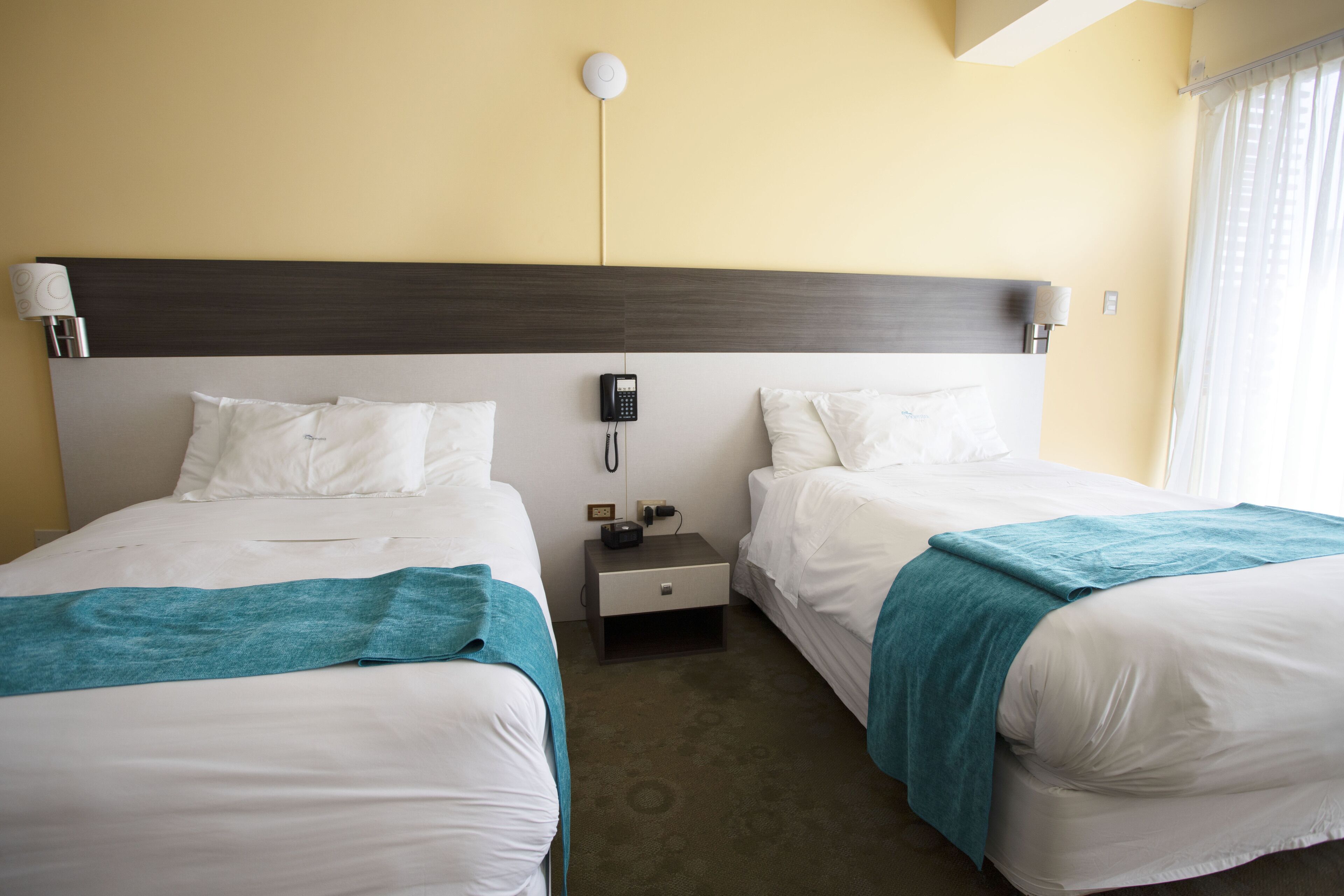 A hotel room with two beds, a nightstand with a phone, and a wall-mounted light fixture.