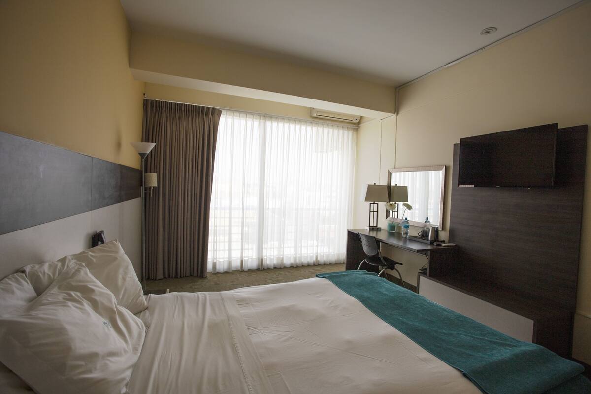 A hotel room with a bed, a desk, a chair, a TV, and a window with curtains.