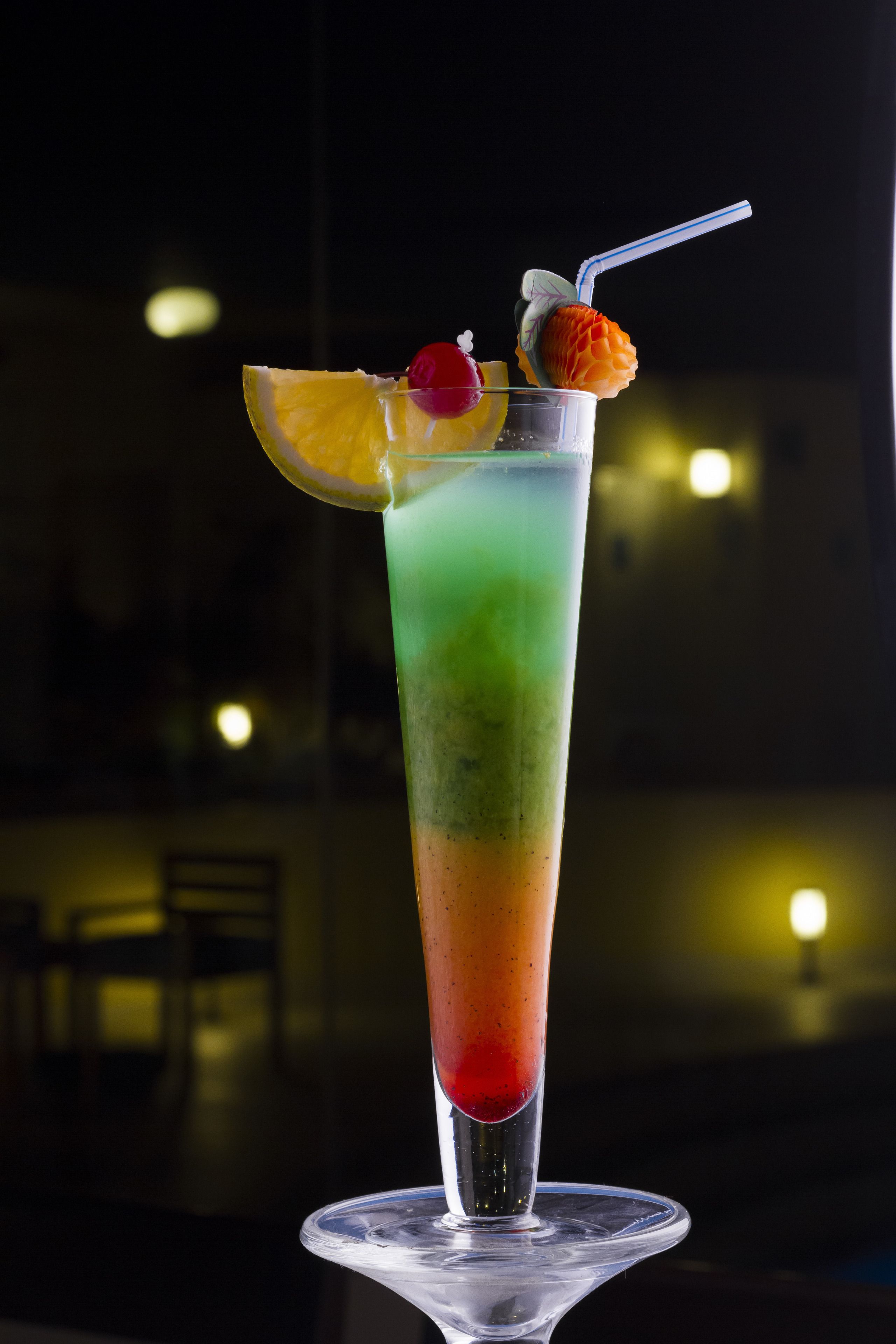 A tall glass with a gradient color drink, garnished with fruit and a straw, on a clear glass base.