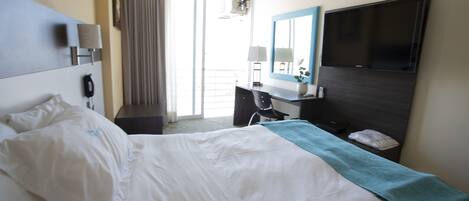 Habitación de hotel con cama, escritorio, silla, televisor y balcón con cortinas.