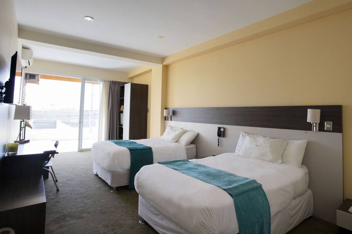 A hotel room with two beds, a desk, a chair, and a view of the outside.