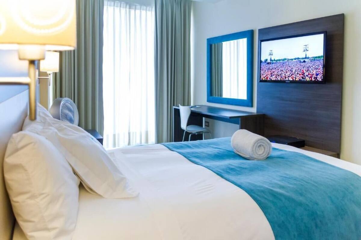 A hotel room with a bed, a desk, a television, and a large window with curtains.