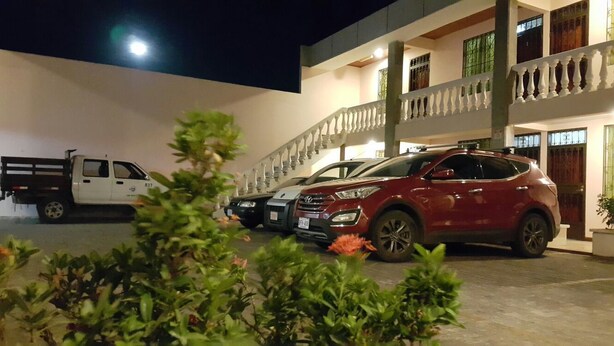 A multi-story building with a white exterior and balconies. There are two cars parked in front, one red and one black. - Hotel Primavera (Liberia)
