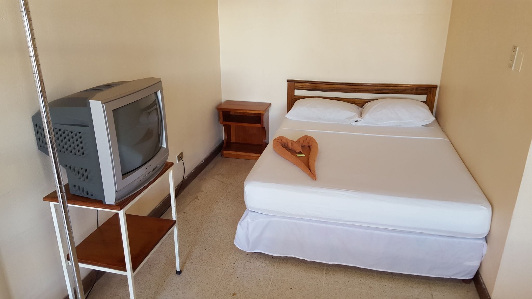 Una cama individual con cabecera de madera, una toalla con forma de corazón y un televisor sobre un mueble.