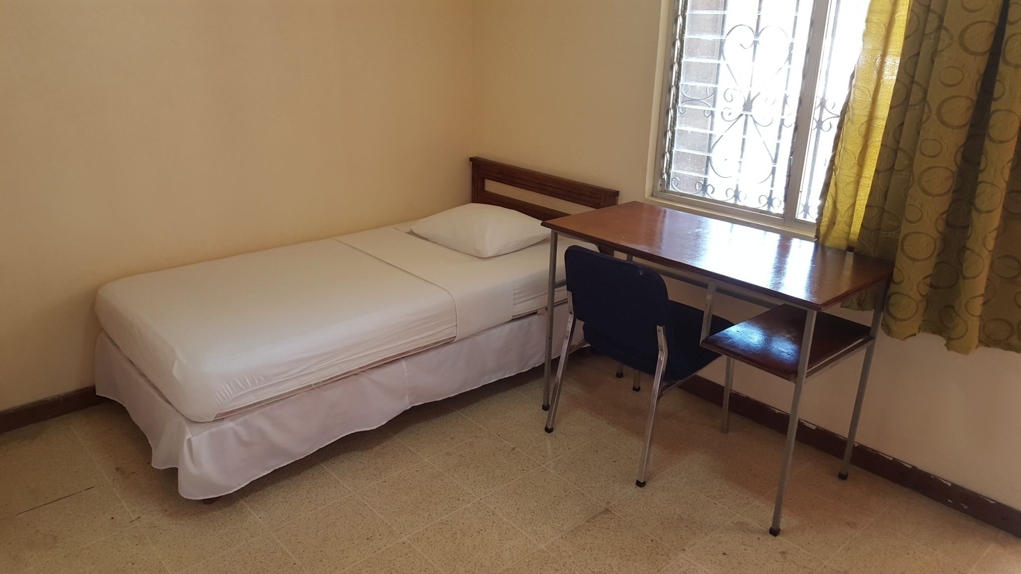Una habitación pequeña con una cama individual, un escritorio y una silla de madera, una ventana con barrotes y una cortina.