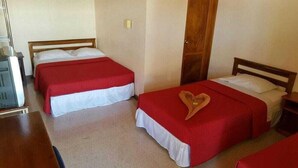 A room with two beds, each with a red bedspread and white pillows. There is a wooden door and a small table with a lamp. - Hotel Primavera (Liberia)