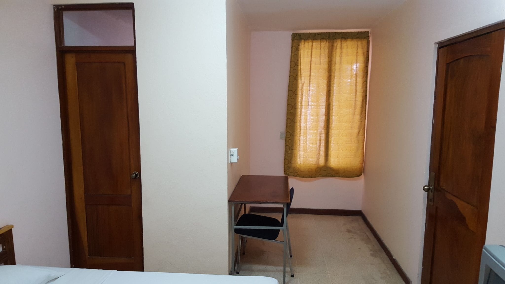 Habitació petita amb una porta de fusta, un escriptori amb una cadira, una finestra amb cortina i un llit.