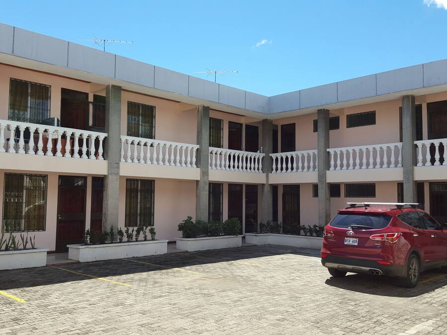 Un edificio de varios pisos con balcones, un coche rojo estacionado adelante y un patio empedrado.