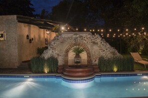 Une piscine agrémentée d’une fontaine en pierre, entourée d’un mur de pierre et illuminée de guirlandes lumineuses.