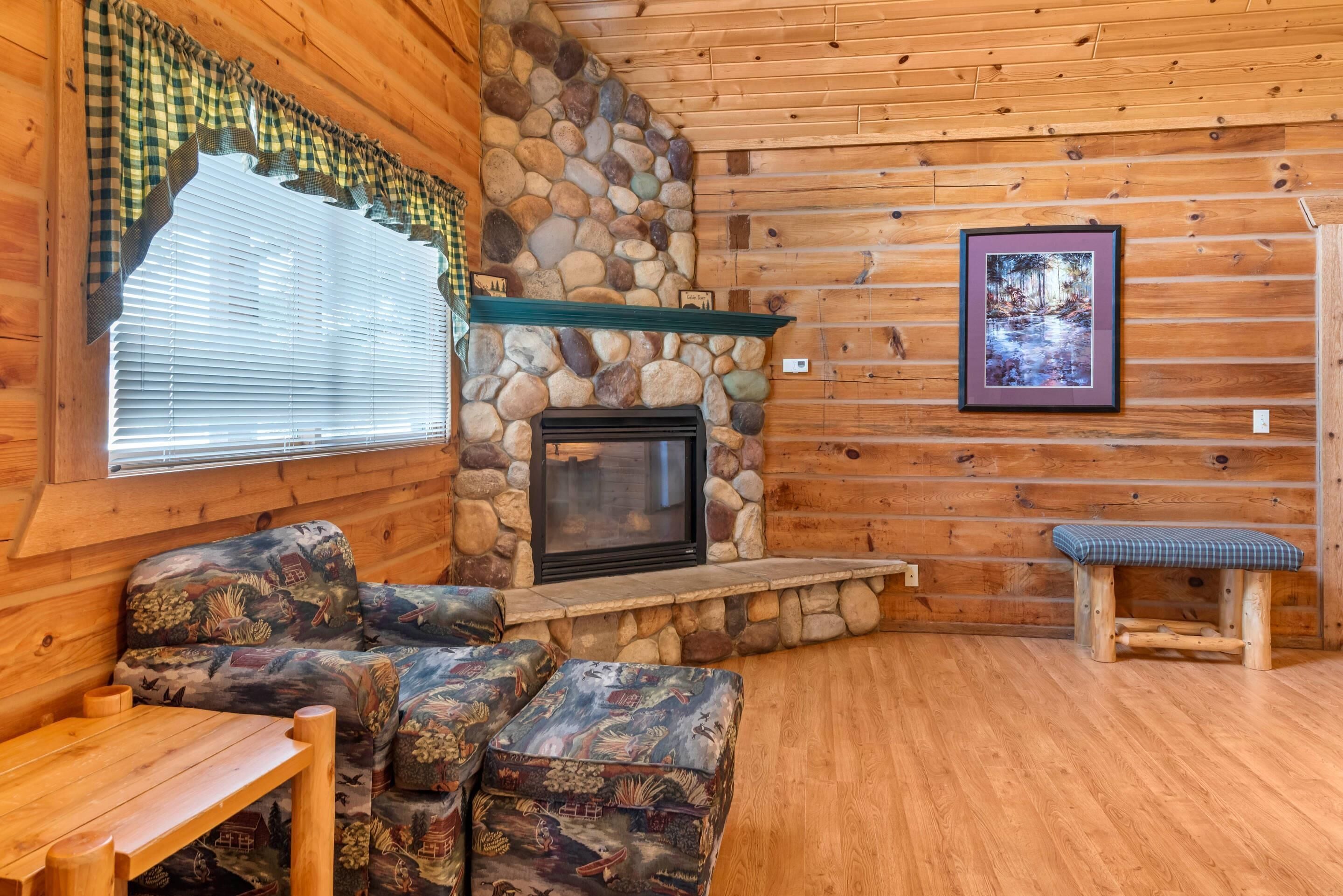 A cozy cabin interior with a stone fireplace, wooden walls, a checkered window valance, a framed picture, and a bench.