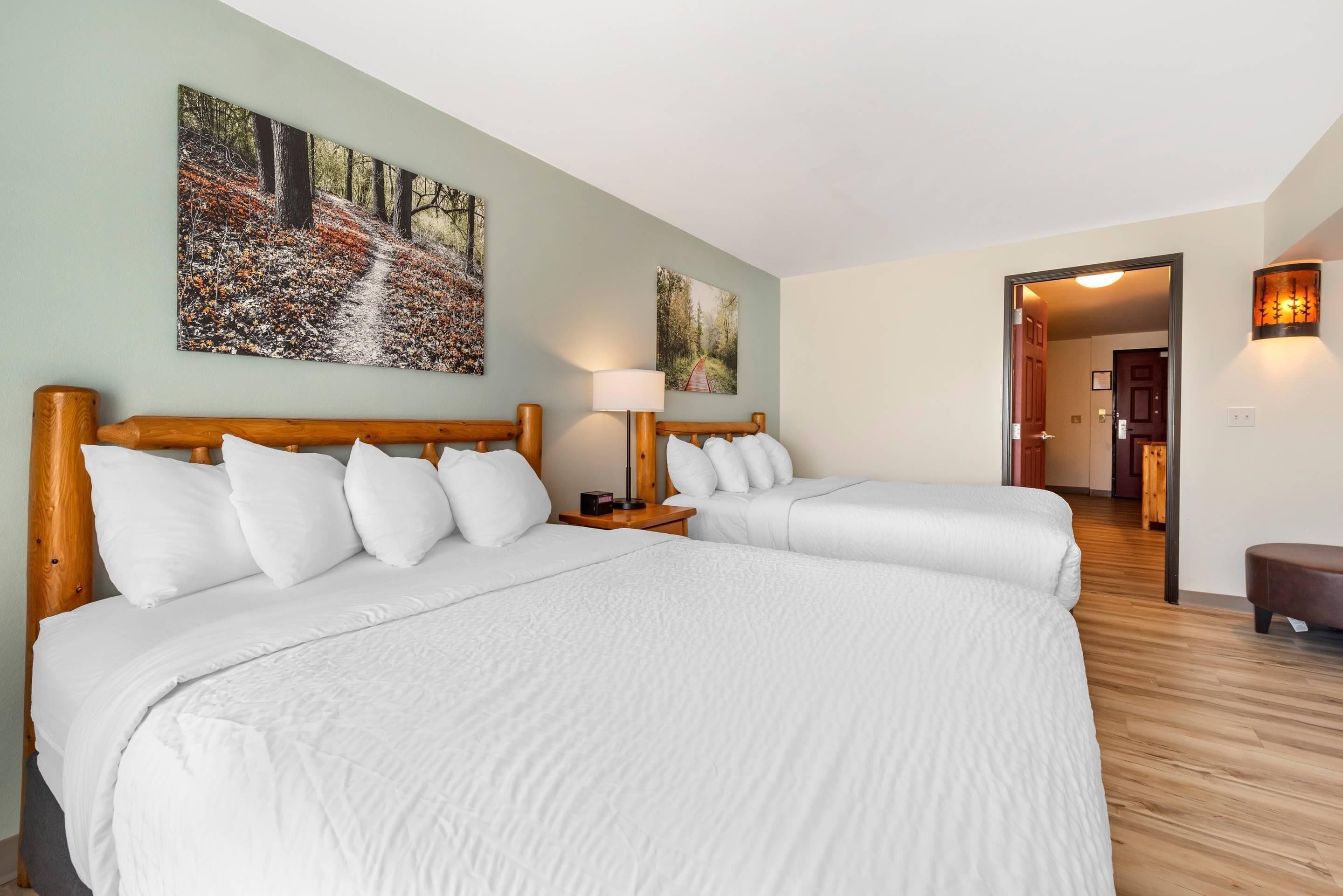 Habitación de hotel con dos camas, cabecero de madera, mesita de noche con lámpara y un cuadro de un sendero en el bosque en la pared.