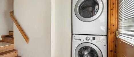 A laundry area with two stacked washing machines, a wooden staircase, and a door with blinds.