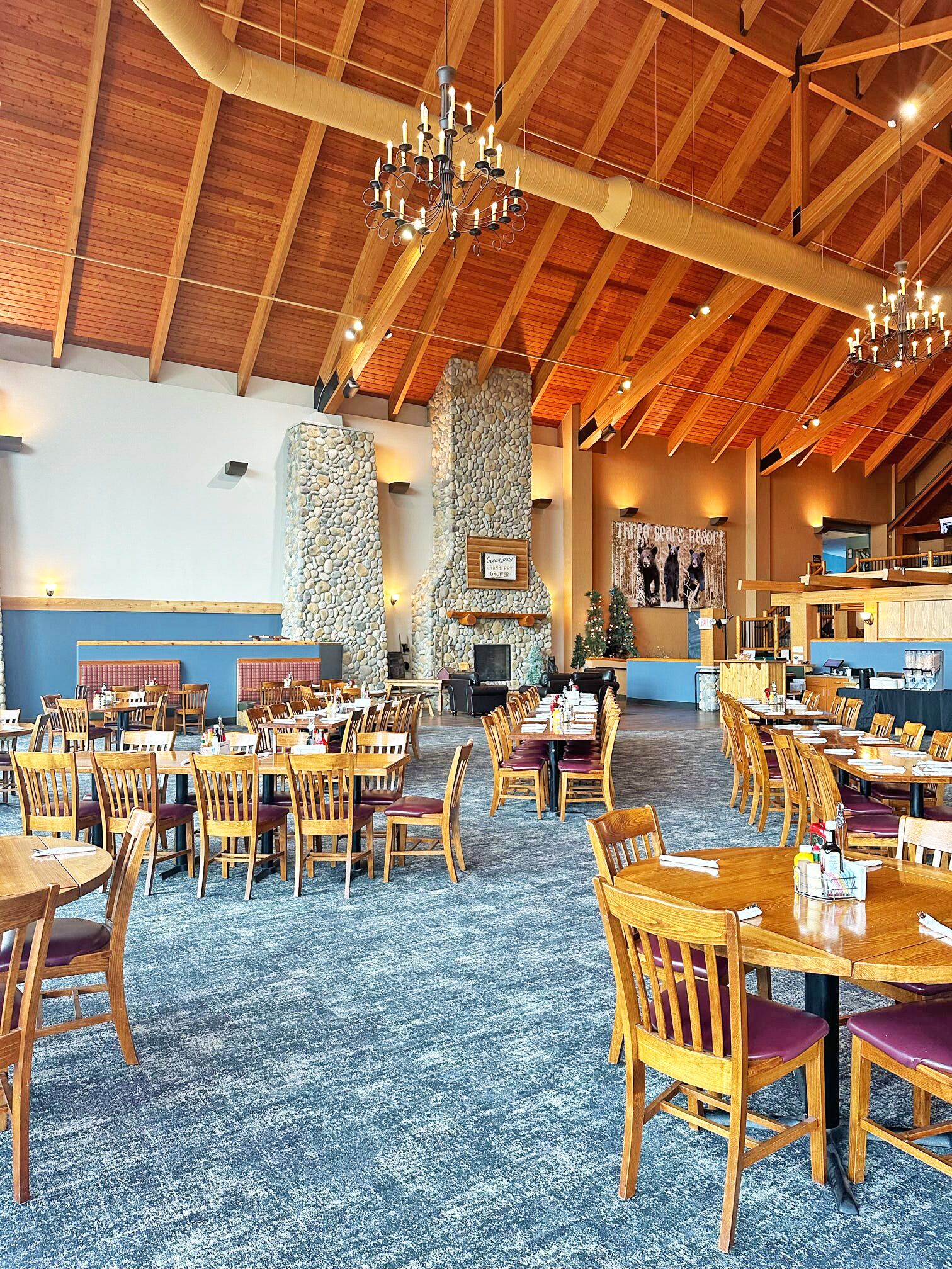 A spacious dining area with wooden tables and chairs, a stone wall, and a high wooden ceiling with chandeliers.
